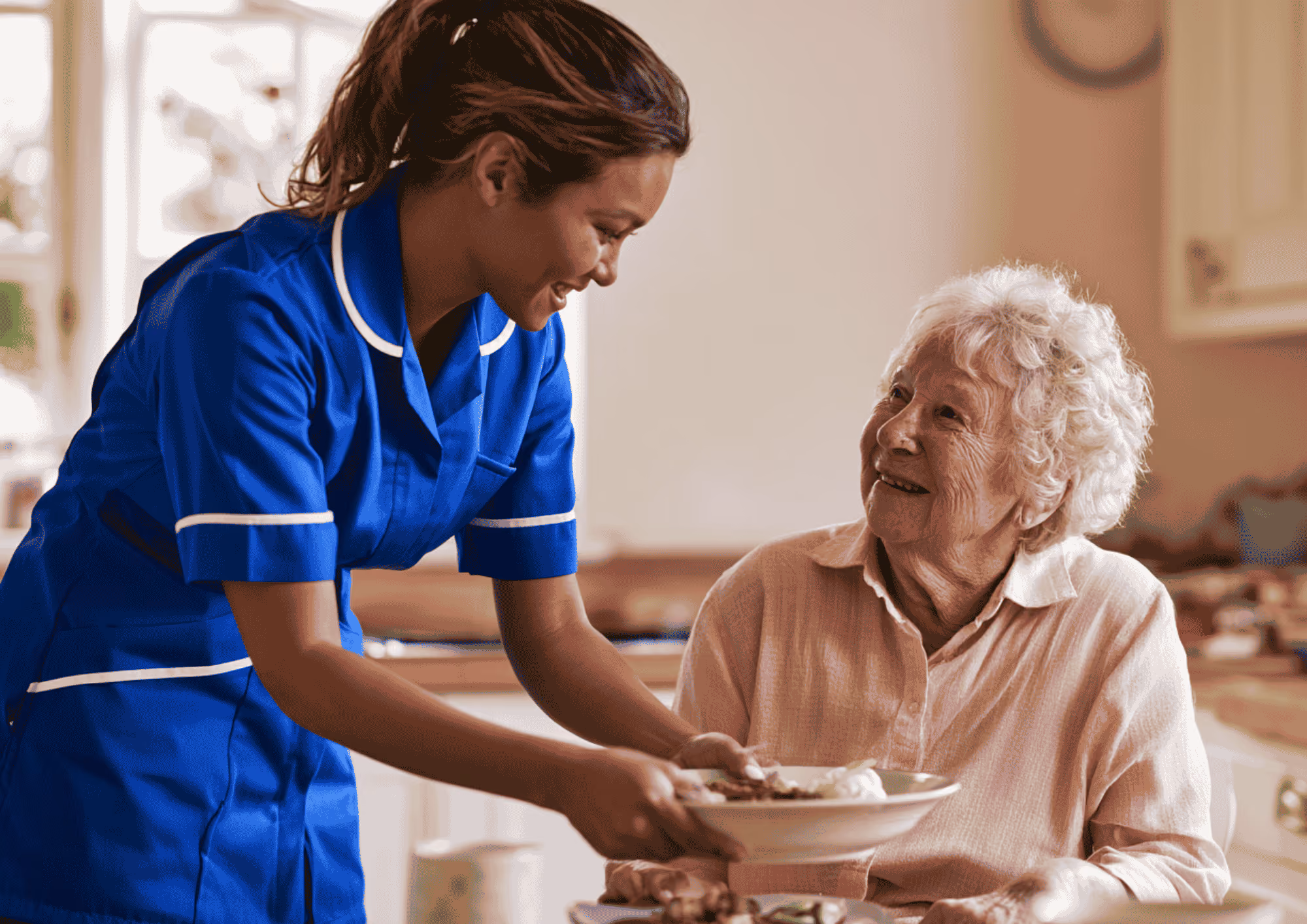 A smiling care worker in a blue uniform serving a meal to an elderly woman at home.