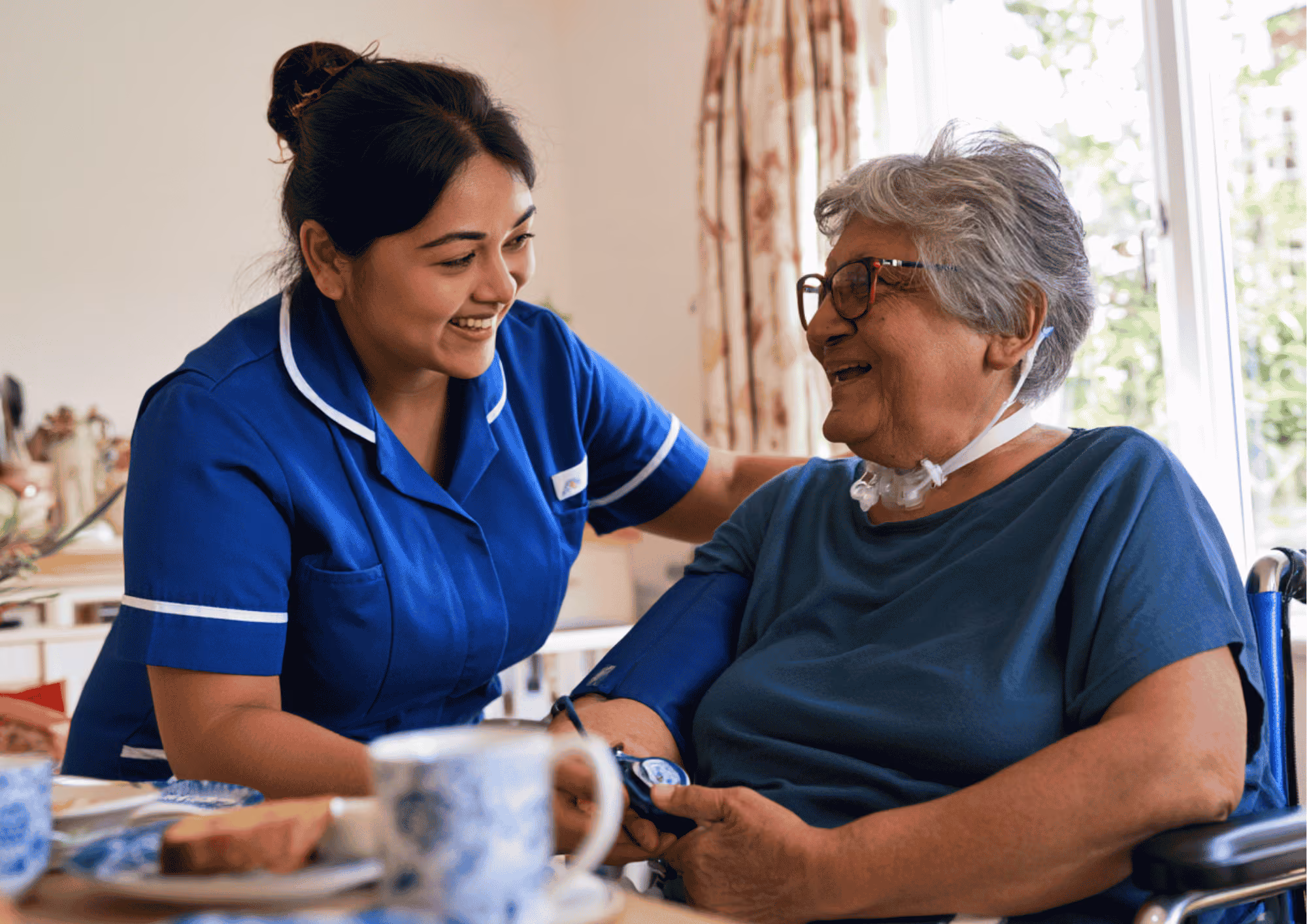 A smiling care worker taking the blood pressure of an elderly woman using a wheelchair at home.