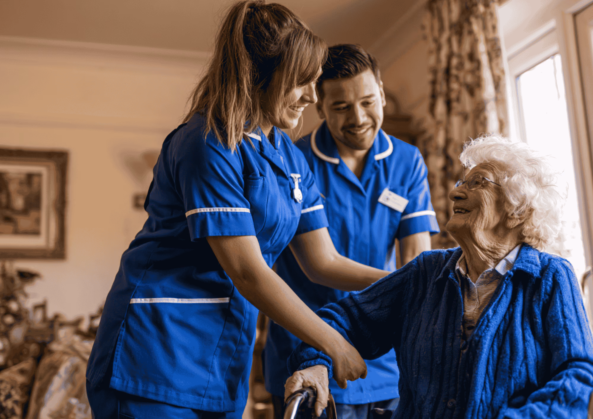Two care workers in blue uniforms supporting an elderly woman as she stands with a mobility frame at home.