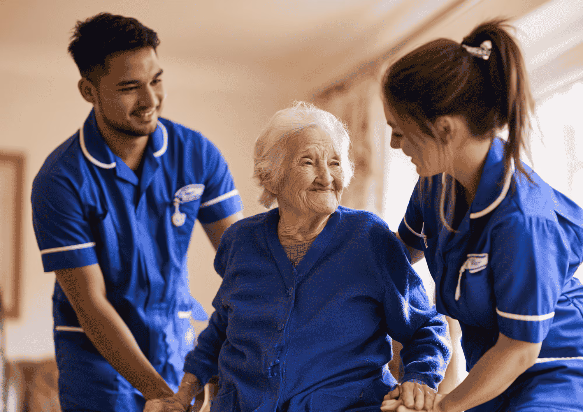 Two care workers in blue uniforms helping an elderly woman rise safely from a chair at home.