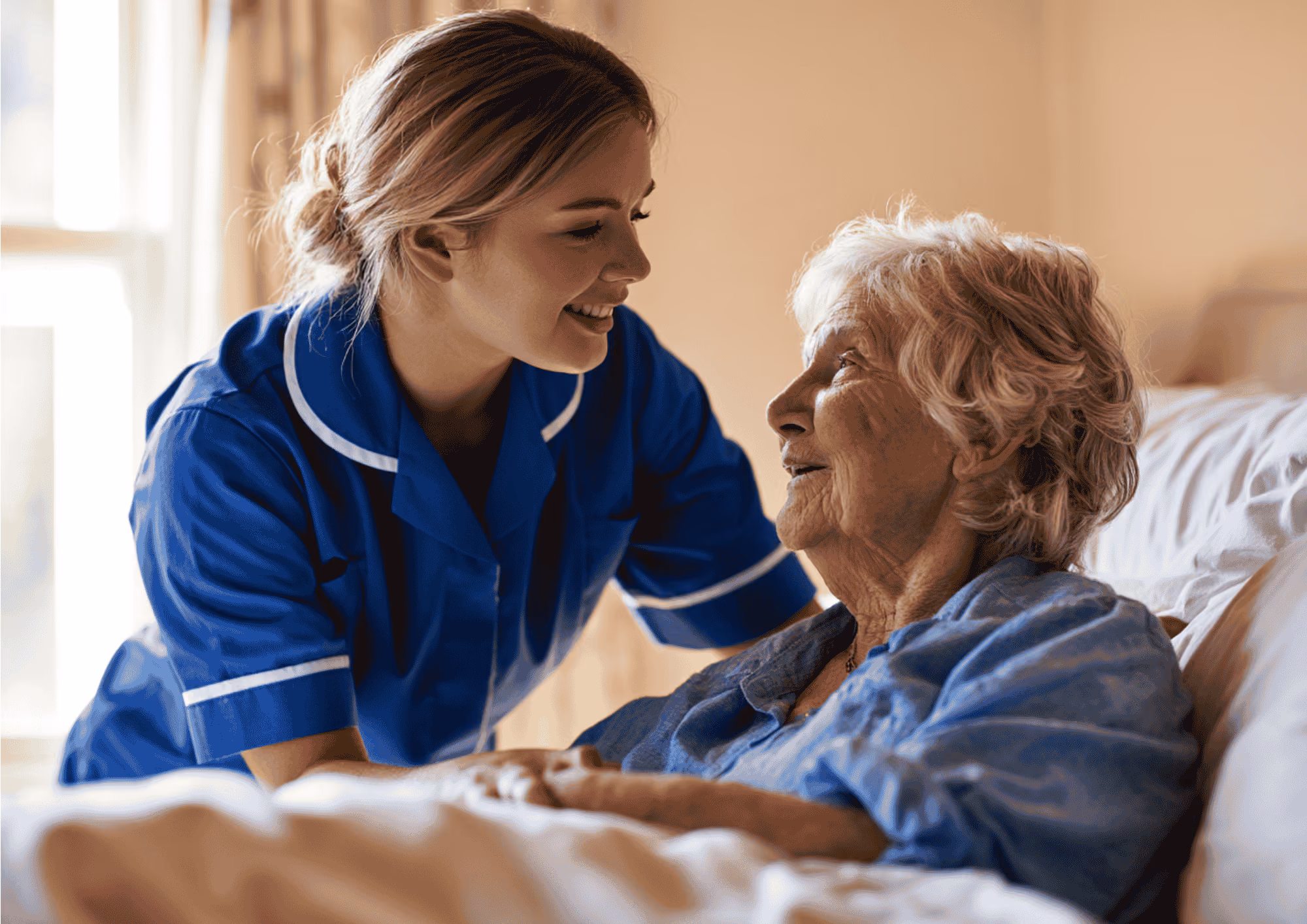 A smiling care worker in a blue uniform helping an elderly woman settle comfortably into bed at home.