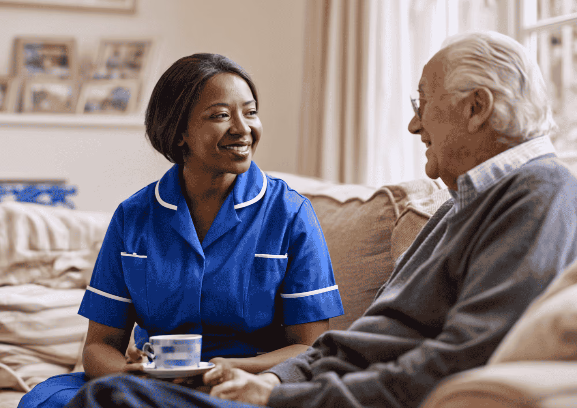 A smiling female caregiver in blue uniform sitting on a couch, holding a cup and saucer, talking with an elderly man wearing glasses and a gray sweater.