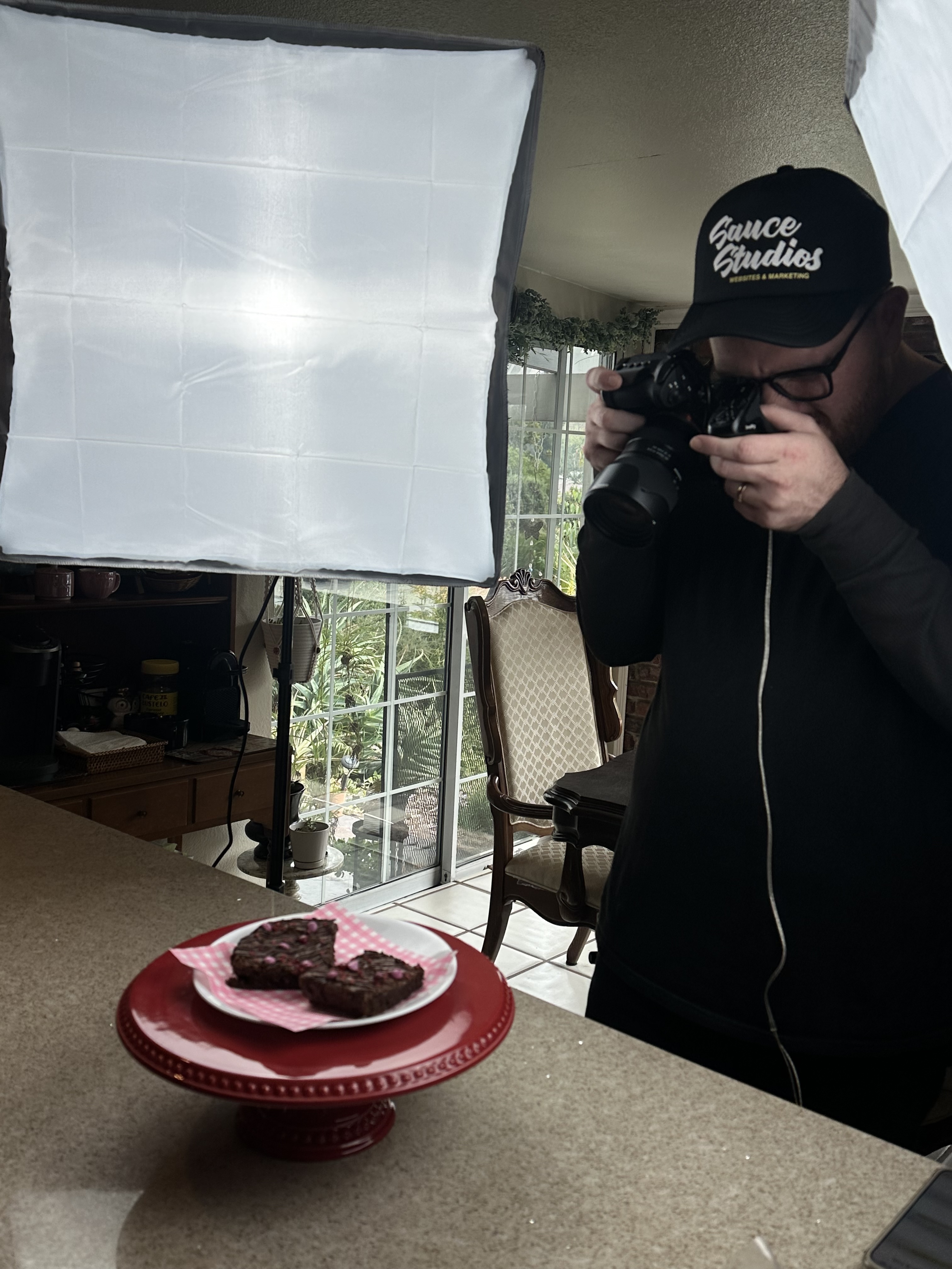 A photographer taking a photo of a man posing with clenched fists near a large softbox light inside a garage with an open door showing the sunset sky outside.