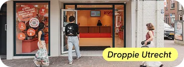 Twee mensen lopen een modern afhaalpunt binnen met een groot oranje bord en een vrouw staat achter de balie, terwijl een derde persoon voorbijloopt buiten op straat.