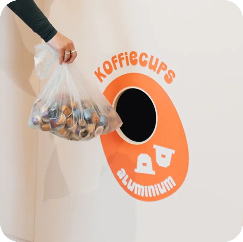 Hand met plastic zak vol gebruikte aluminium koffiecapsules vlakbij een oranje ophaalpunt op een witte muur met de tekst Koffiecups aluminium.