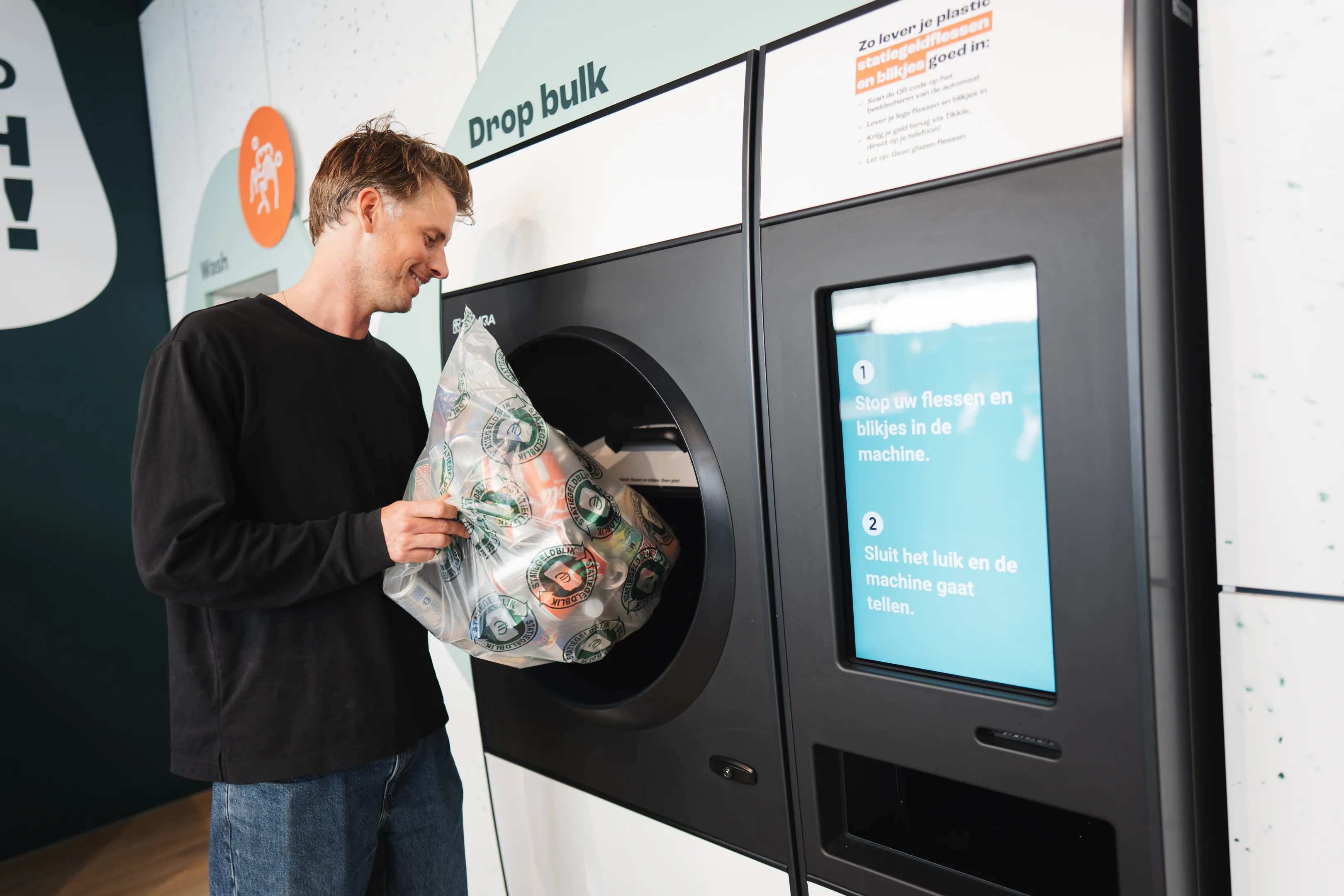 Man in zwarte trui die een doorzichtige plastic zak met lege flessen en blikjes in een recycleermachine met scherm bij een Drop bulk-locatie stopt.