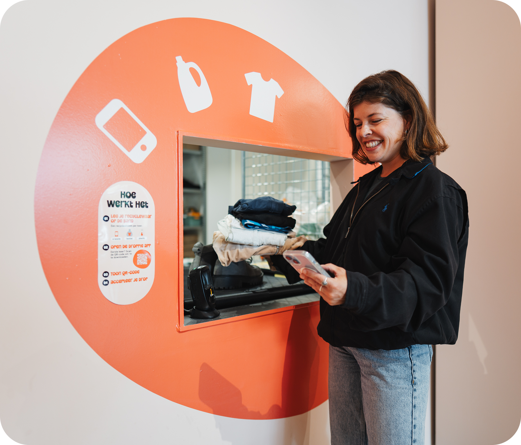 Bij Droppie: Vrouw die glimlacht terwijl ze een stapel gevouwen kleding vasthoudt en haar telefoon gebruikt voor een oranje recyclagemachine met pictogrammen van een fles, een t-shirt en een mobiel.