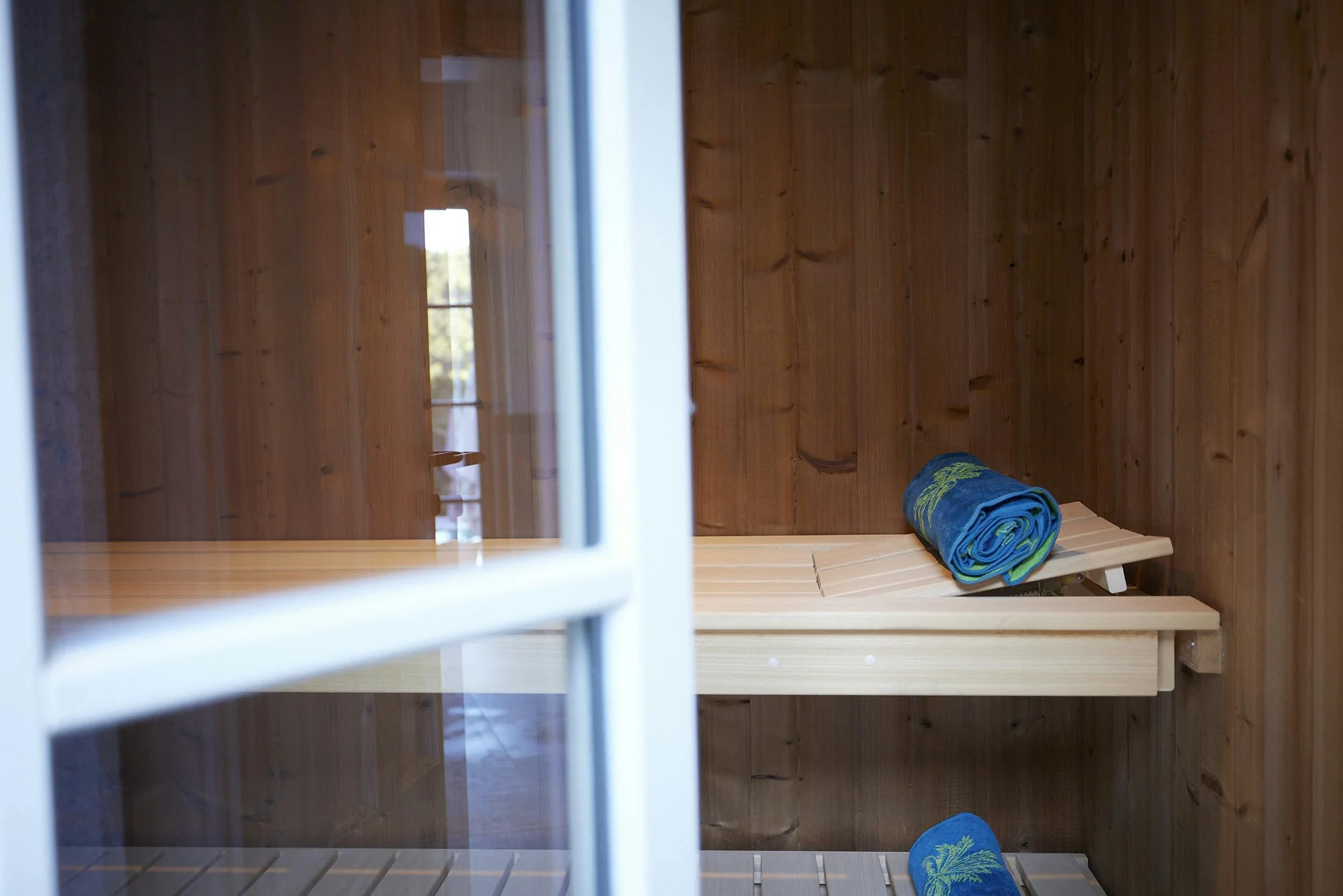 Detail of the wooden sauna interior with a bench and rolled towel, visible through a glass door. ByMilagro's luxury amenities.