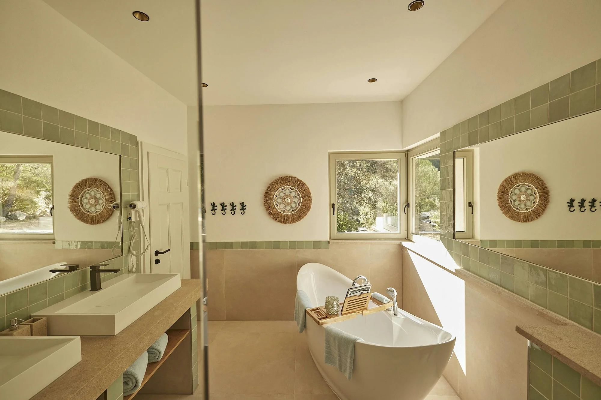 Ensuite luxury bathroom with freestanding bathtub, double vanity, green accent tiles, and window with natural views. Premium ByMilagro accommodation.