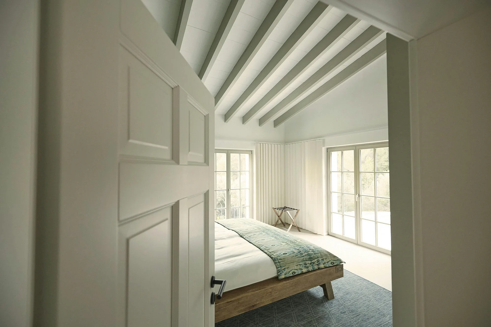 Bright, airy luxury bedroom view through an open door, featuring a high vaulted ceiling with grey beams and double doors leading to a balcony. ByMilagro rental.