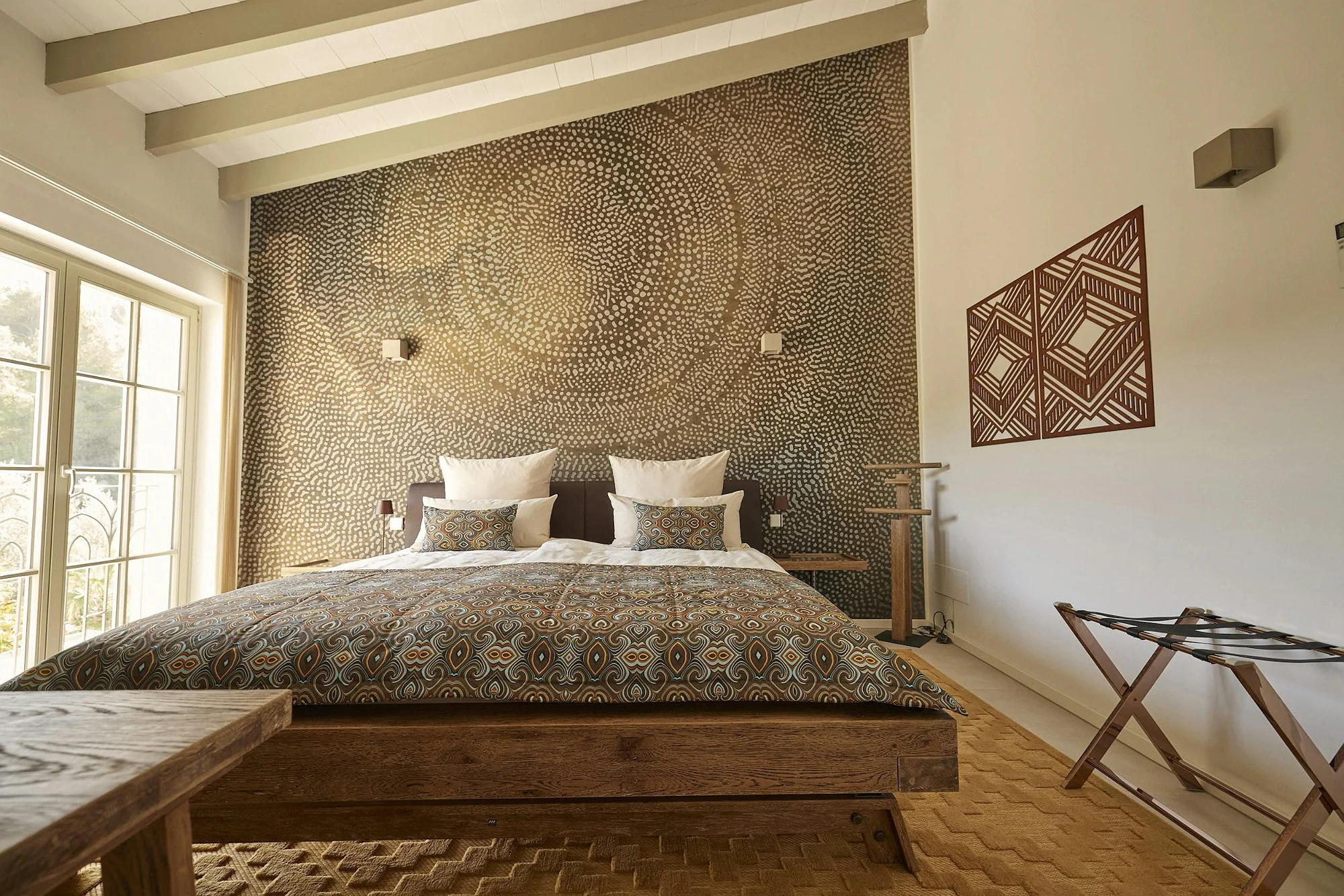 ByMilagro bedroom with wooden bed frame, patterned textiles, textured golden accent wall, and high ceilings with exposed beams.