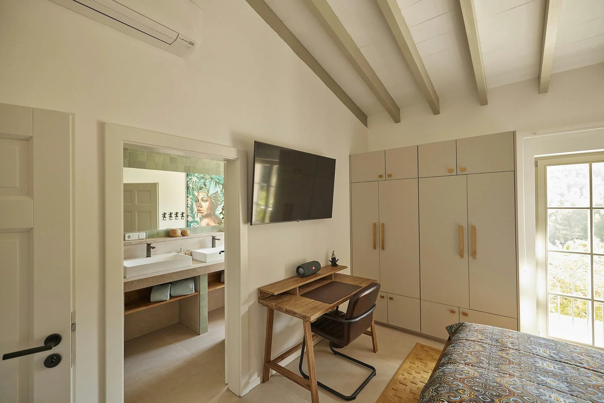 Modern ByMilagro bedroom with built-in wardrobe, wooden work desk, wall-mounted TV, and an open doorway leading to a stylish ensuite bathroom.