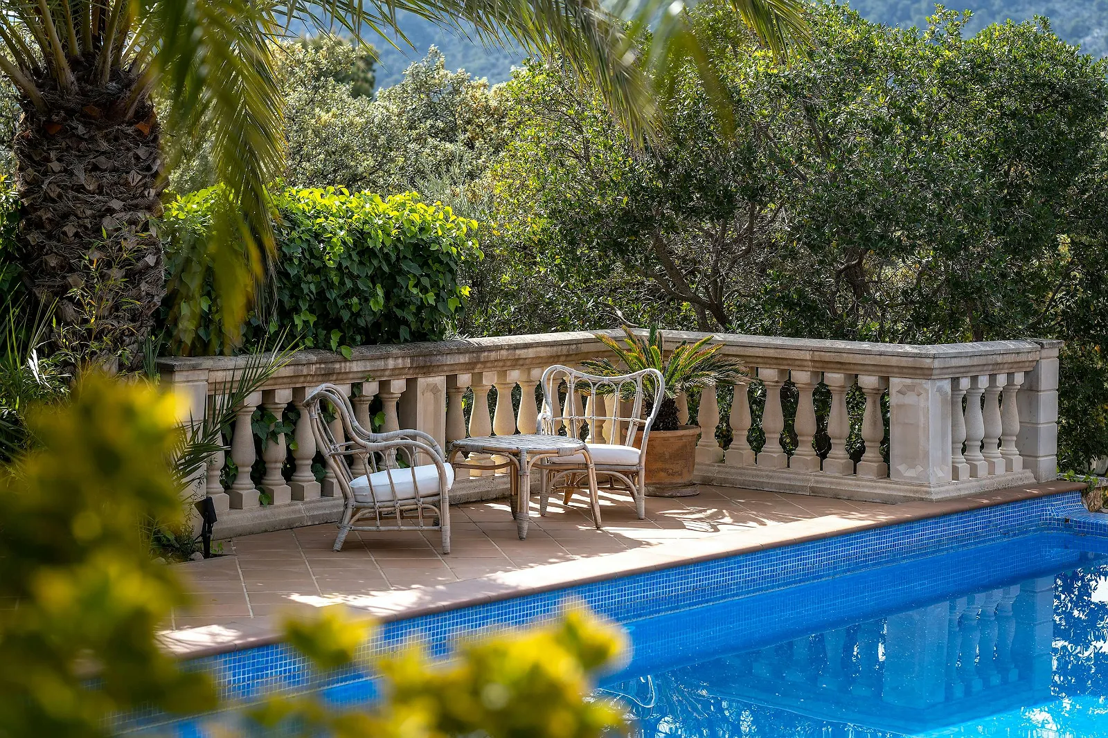 Poolside seating area with antique wicker chairs and table next to the stone balustrade, overlooking the water at Villa Scarlett luxury rental.
