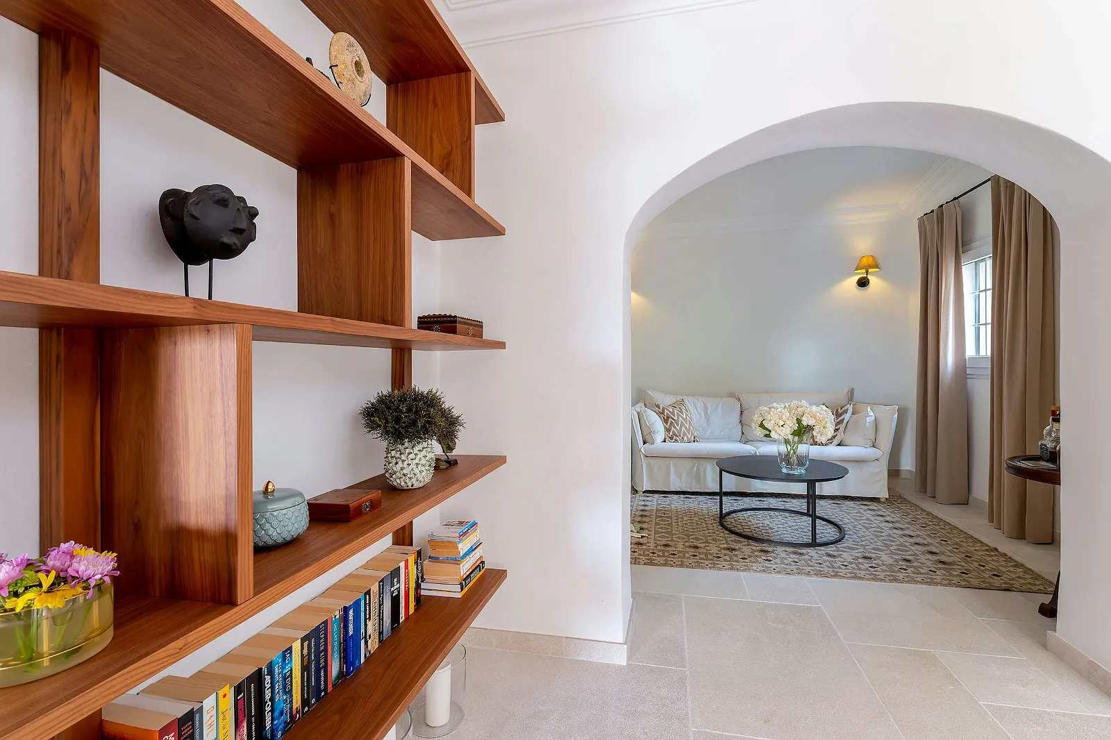 Interior hallway with modern wooden bookshelves and an archway leading into the bright, white-walled living room. Elegant interior design ByMilagro.