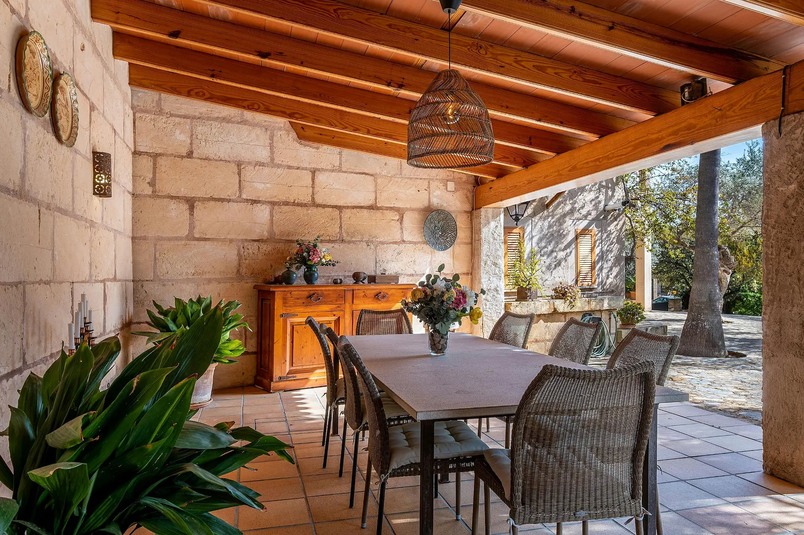 Covered outdoor dining area at Villa Scarlett with stone walls, wicker chairs, wooden sideboard, and a large dining table. Al fresco dining ByMilagro.