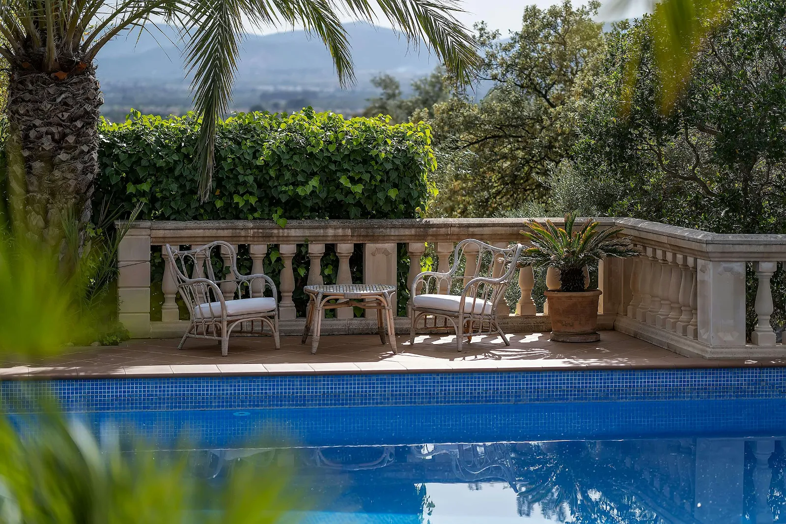 Romantic seating area by the swimming pool with white cushions and a backdrop of lush greenery and distant mountains at ByMilagro's Villa Scarlett.
