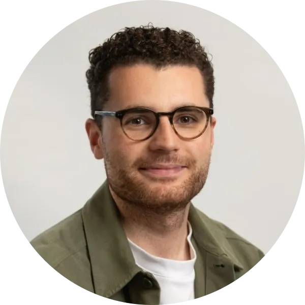 Portrait of Thomas Gorst wearing brown circular glasses, a forest green shirt over a white T-shirt, against a soft grey background.