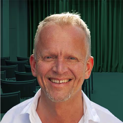 Leende man med kort blont hår och skägg i en vit skjorta framför gröna gardiner och stolar.