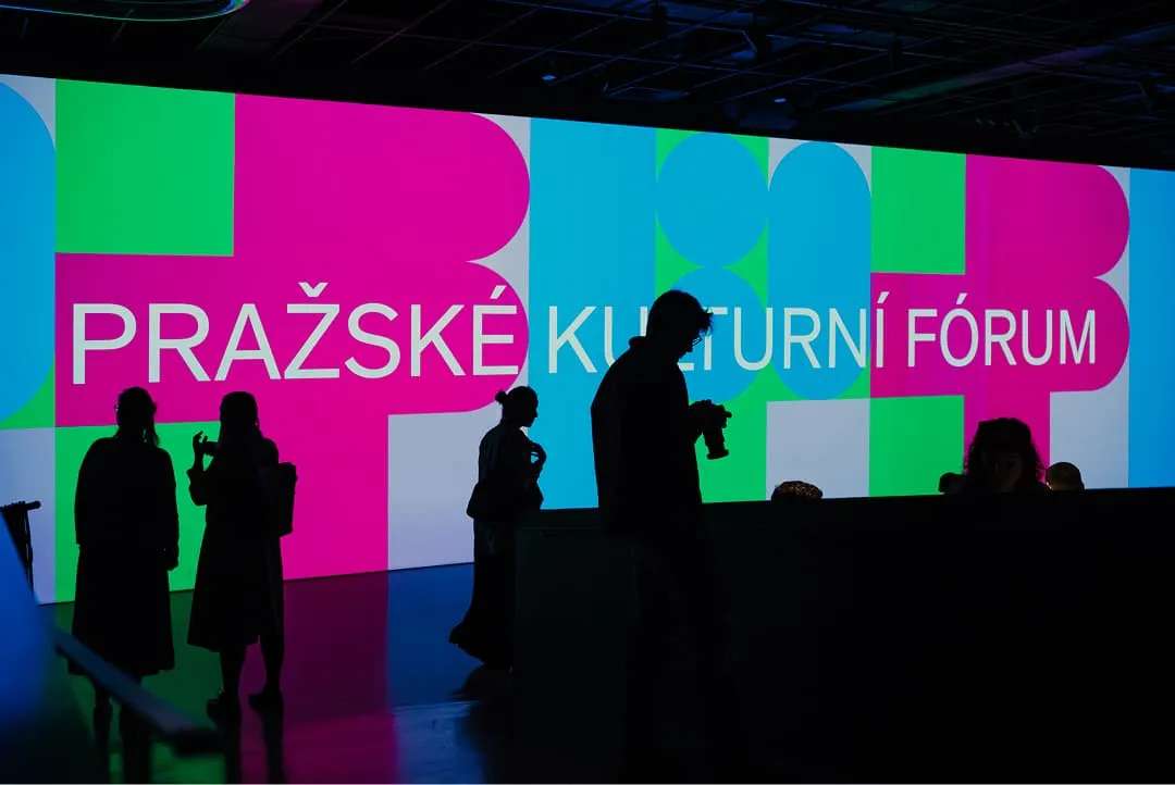 Siluety lidí stojících před velkou barevnou digitální obrazovkou, na které je zobrazen text „PRAŽSKÉ KULTURNÍ FÓRUM“.