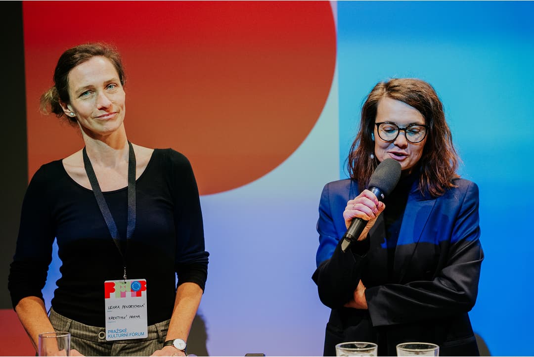 Dvě ženy stojící před barevným pozadím; jedna žena mluví do mikrofonu, zatímco druhá poslouchá.