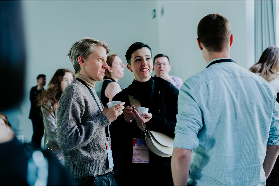 Skupina lidí zapojená do konverzace, dvě ženy s kávovými šálky v rukou a s odznaky akce.
