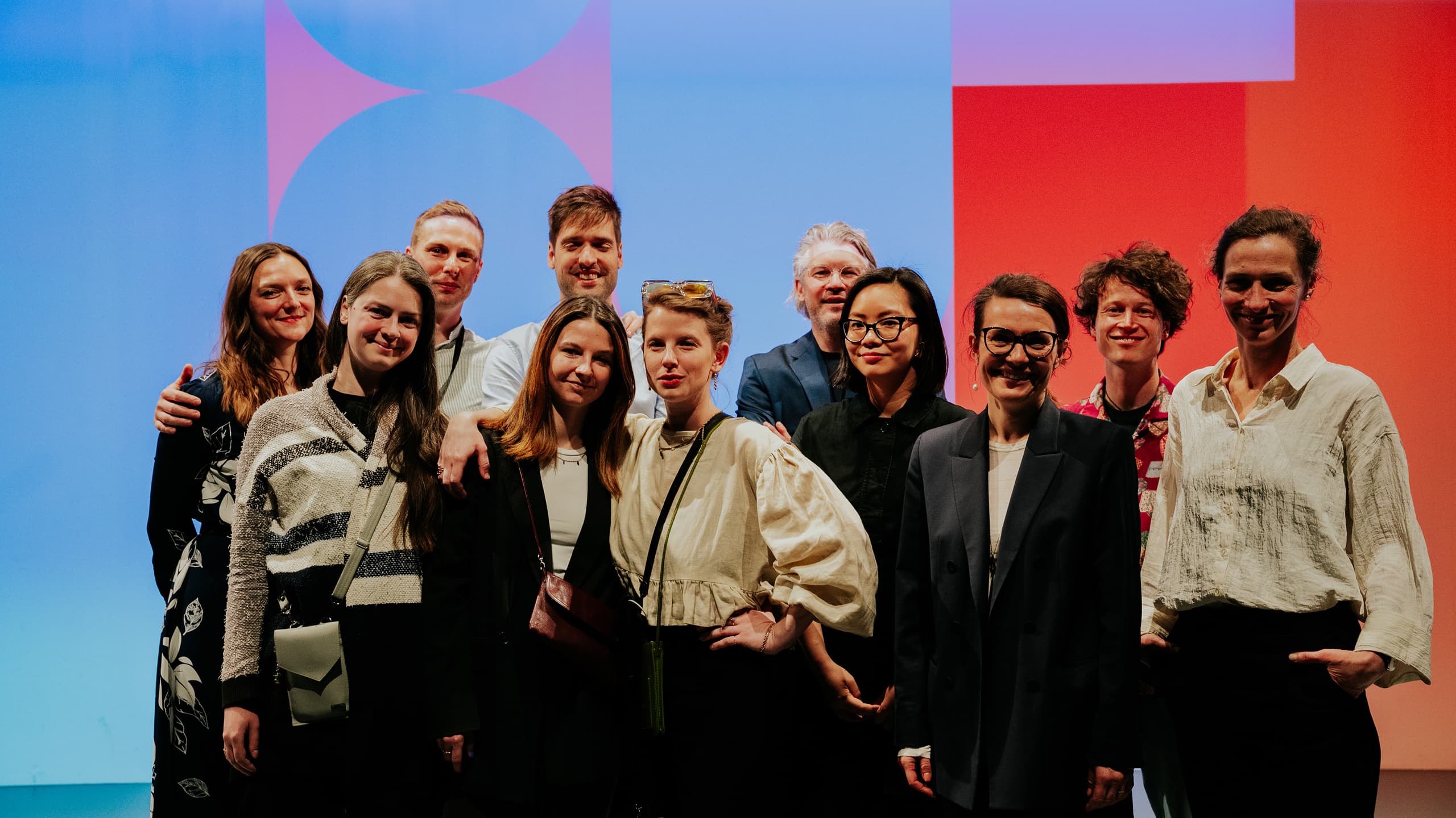 Skupinová fotografie jedenácti usmívajících se lidí stojících těsně vedle sebe před barevným modro-červeným pozadím.
