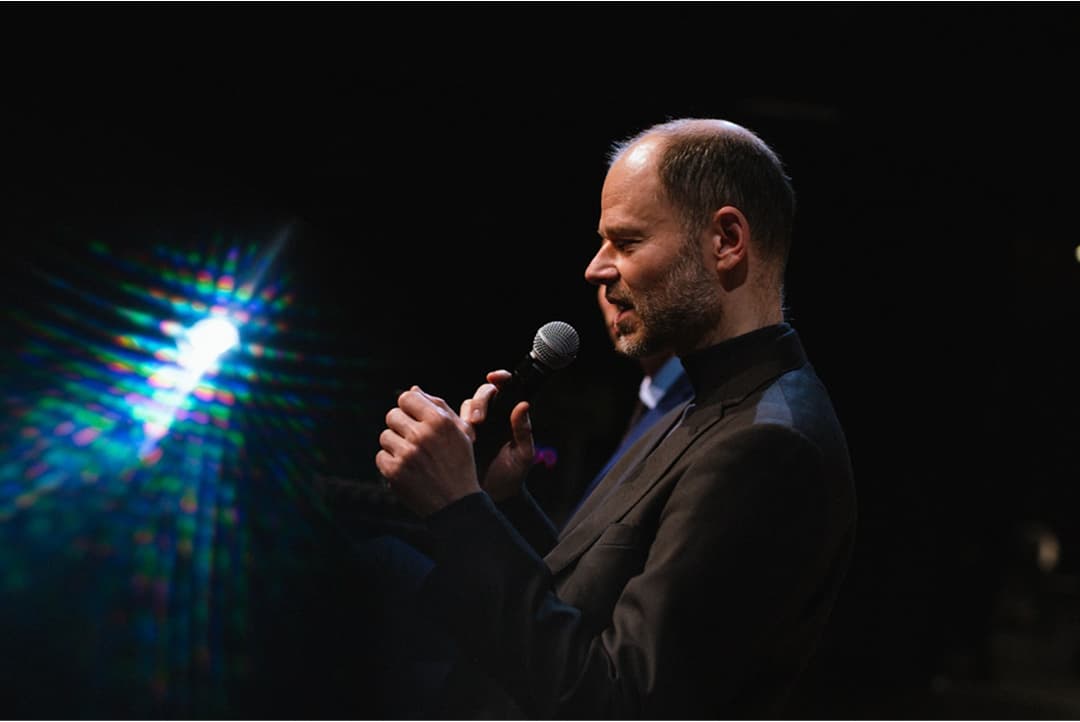 Muž v tmavém obleku drží mikrofon a mluví před černým pozadím s jasným světlem a barevným odleskem vlevo.