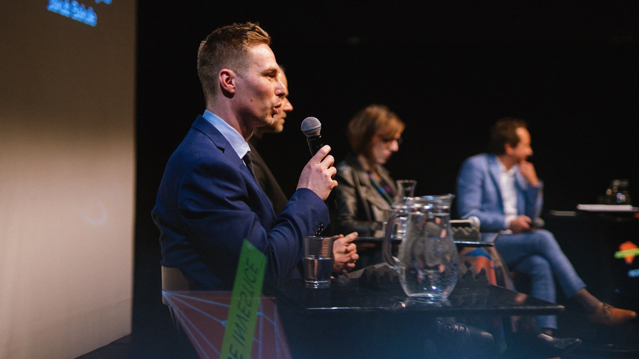 Muž v modrém obleku hovoří do mikrofonu během panelové diskuze se třemi dalšími sedícími účastníky v tlumeně osvětlené místnosti.