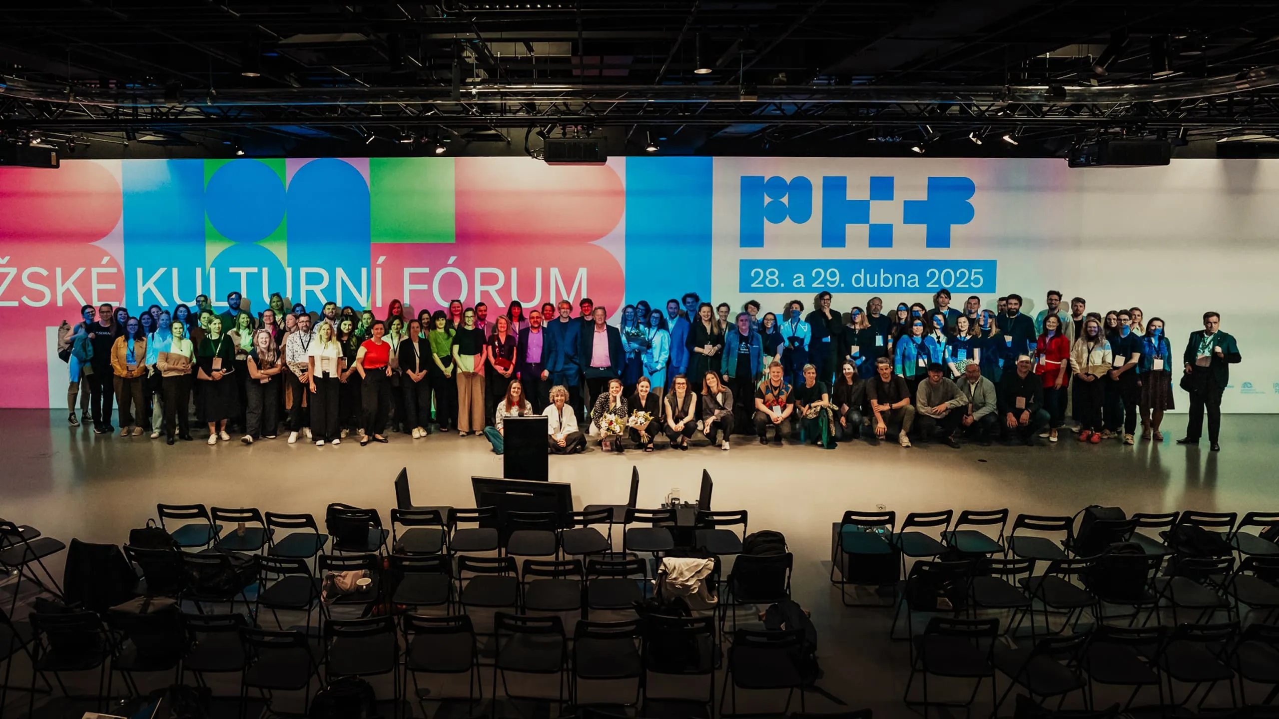 Velká skupina lidí pózuje pro fotografii před barevným pozadím s nápisem „KULTURNÍ FÓRUM“ a daty akce „28. a 29. dubna 2025“.