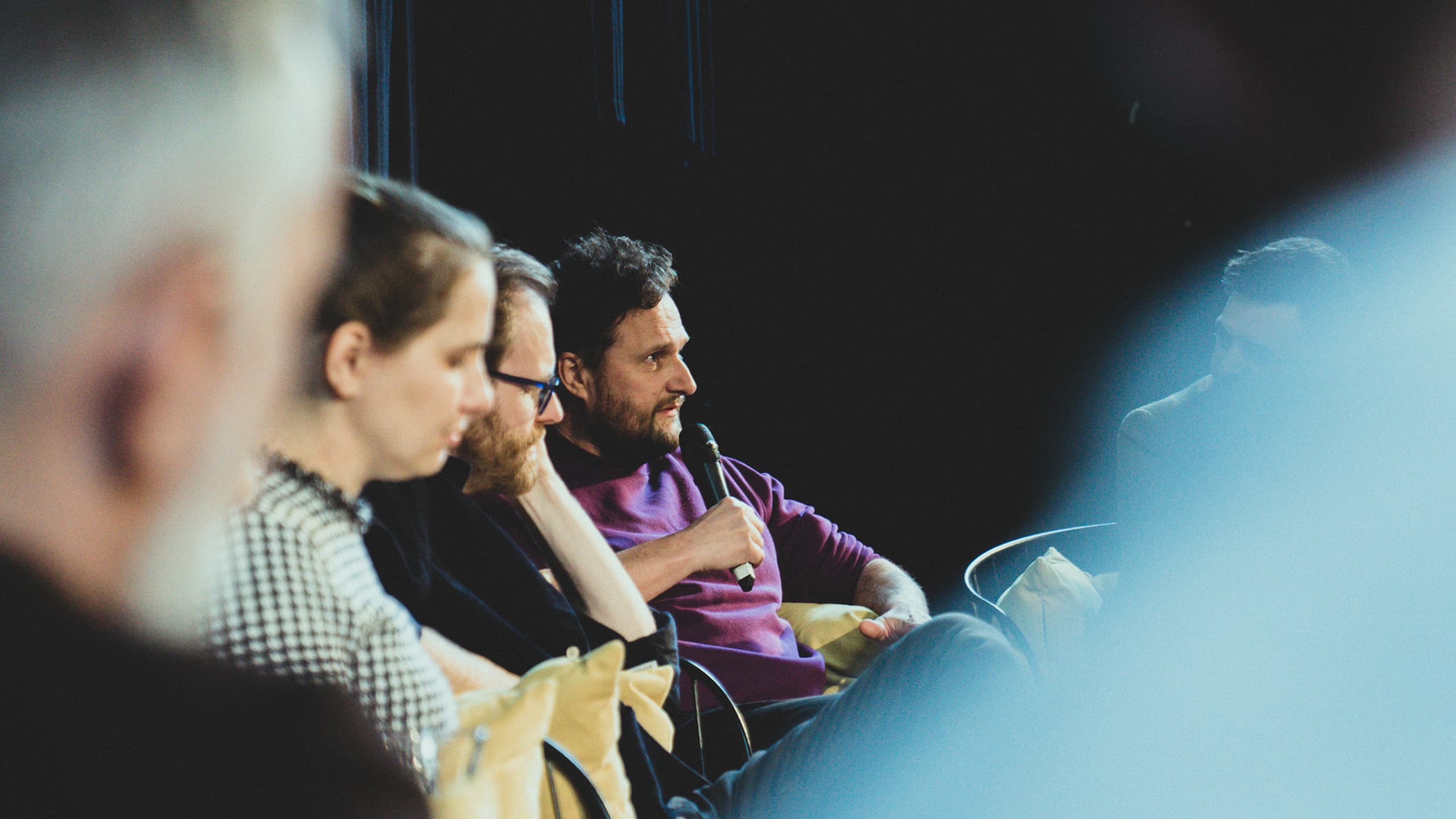 Muž v fialovém svetru mluví do mikrofonu během panelové diskuze se třemi dalšími lidmi sedícími vedle něj.