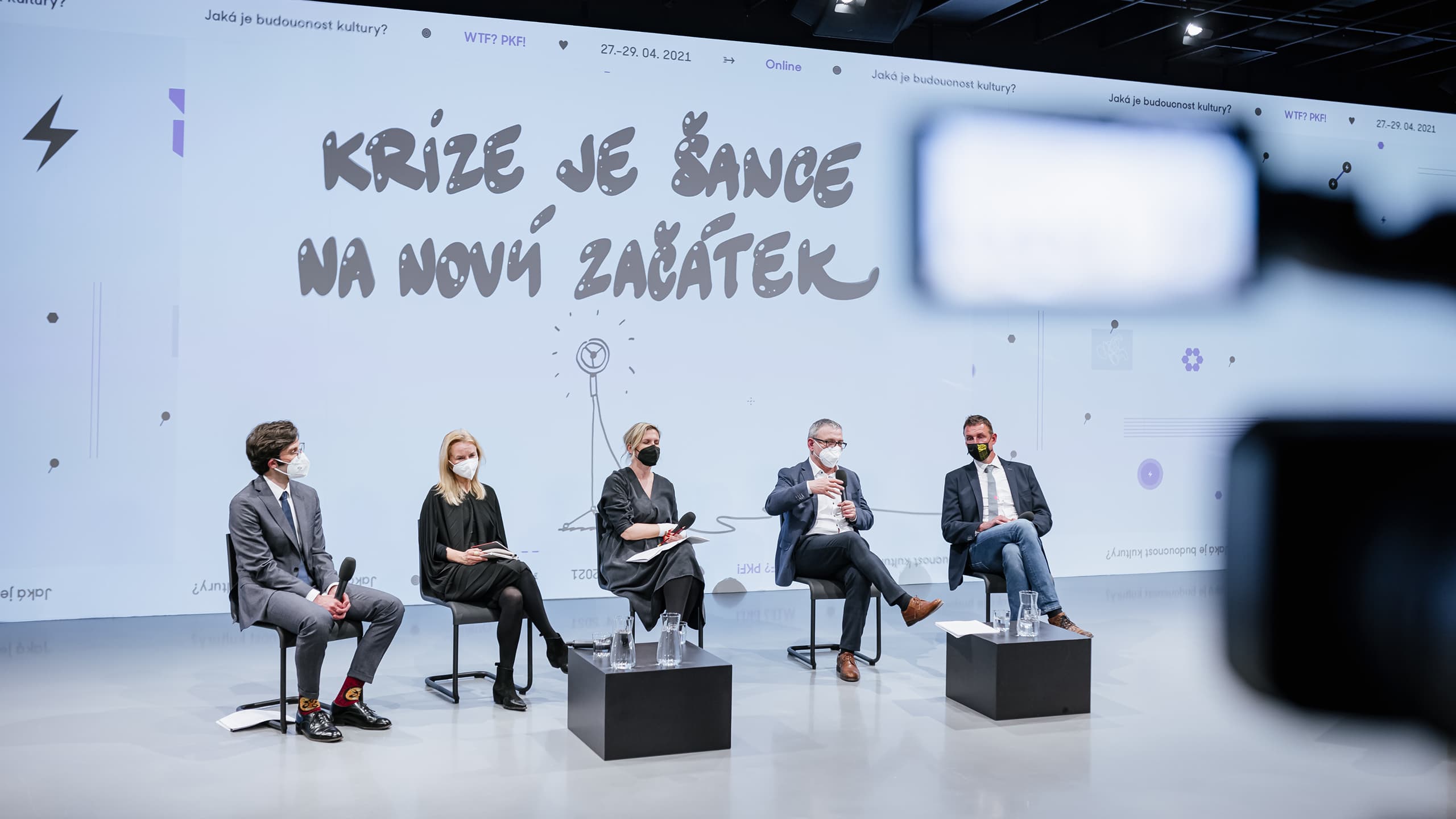 Pět panelistů s rouškami na obličeji sedí na židlích během diskuse na pódiu s velkým bílým pozadím, na kterém je český text a grafika.