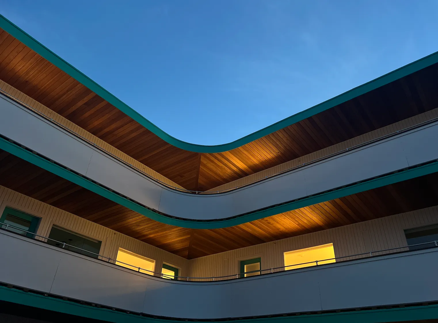 View looking up at a modern two-story building with wooden ceilings and glowing yellow balcony windows against a clear evening sky.