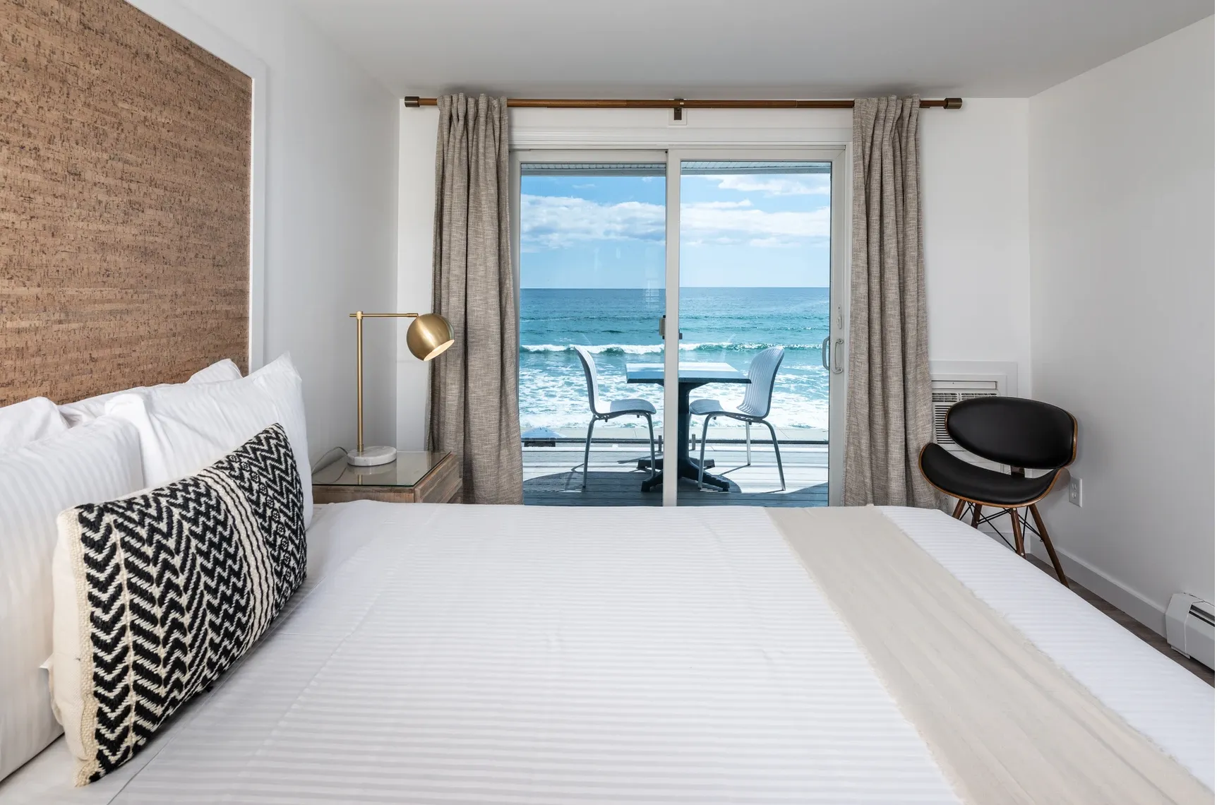 Bedroom with a large white bed facing sliding glass doors that open to a balcony with two chairs and a table overlooking the ocean.
