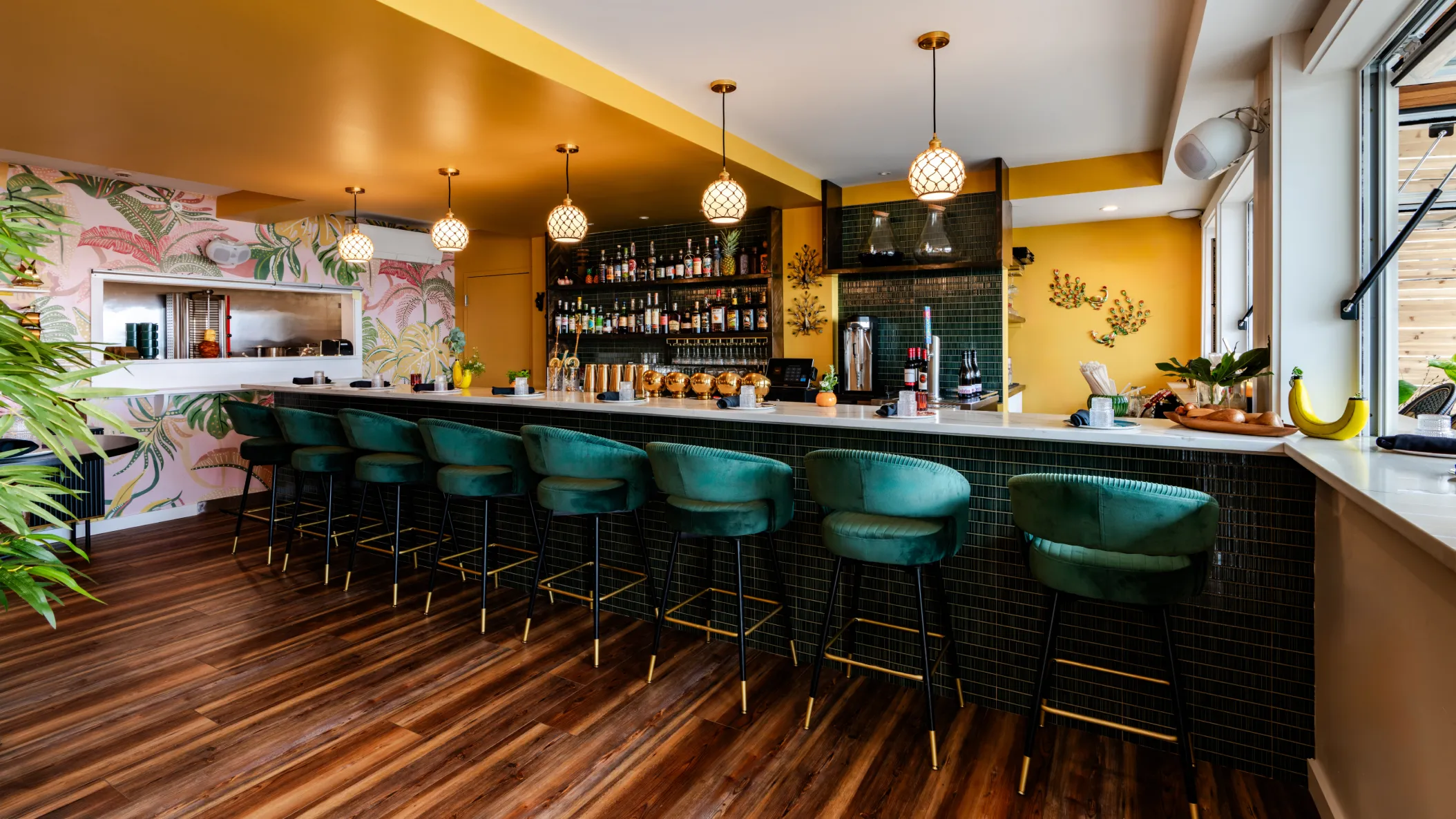 Bright modern bar with green velvet stools, tropical wallpaper, gold accents, and pendant lighting.
