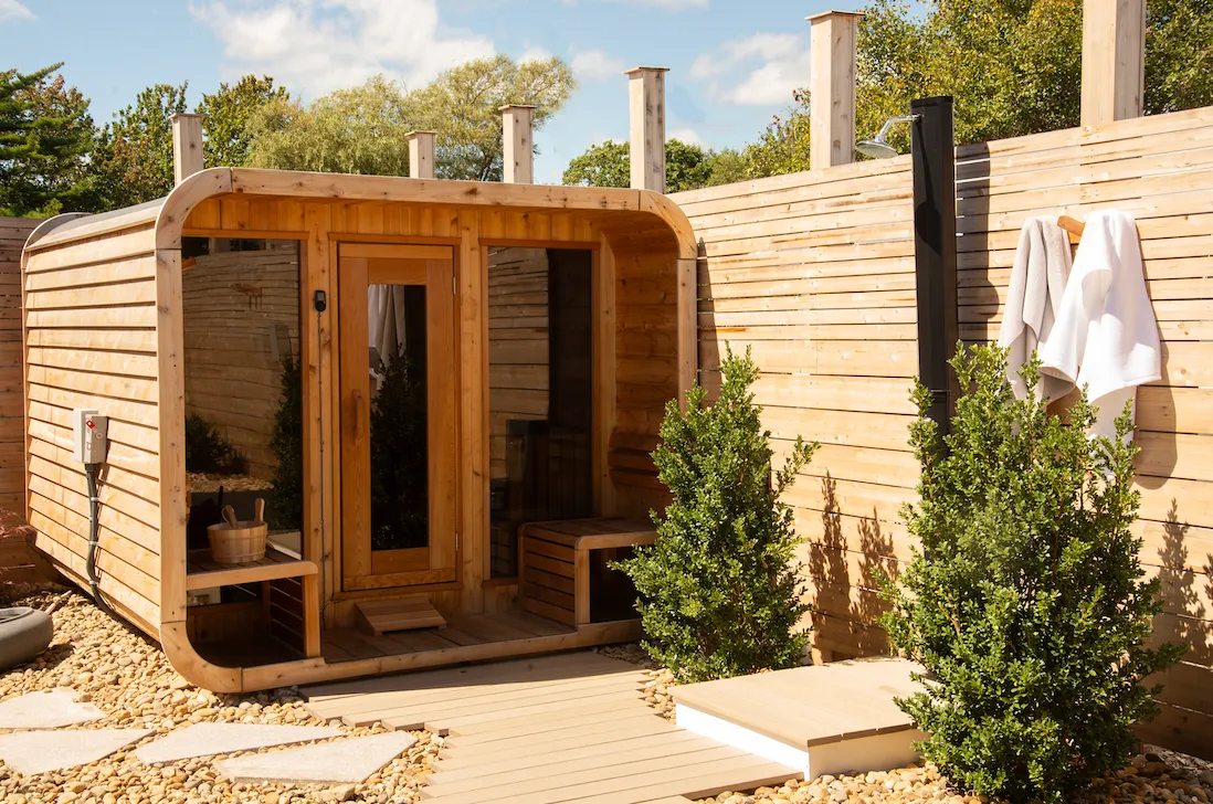 Outdoor wooden sauna cabin with glass door, adjacent wooden wall with towels hanging and green shrubs around.