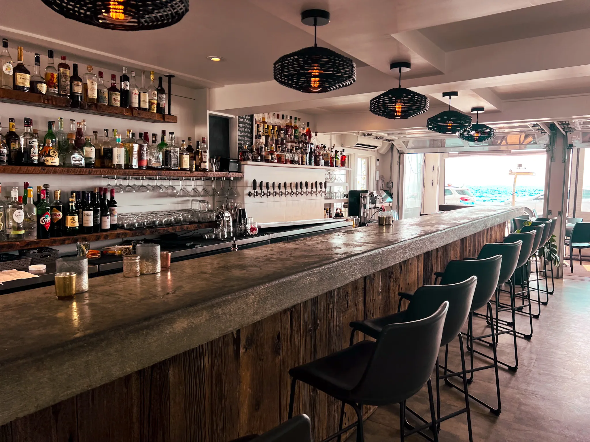 Interior of a modern bar with wood and concrete counter, lined with black bar stools, shelves of liquor bottles, and pendant lights overhead.