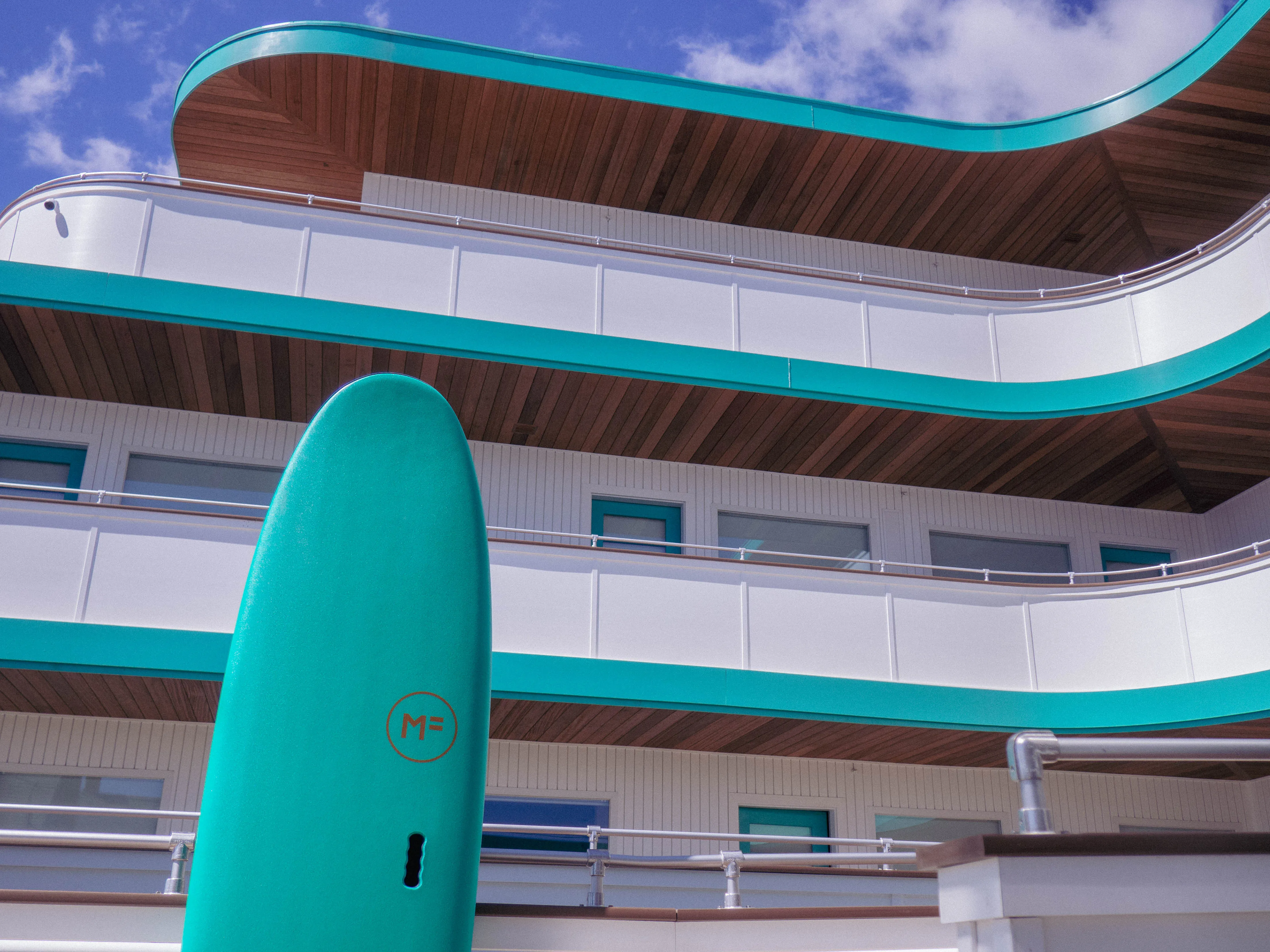 Turquoise surfboard standing upright in front of a modern building with curved balconies and wooden ceilings under a partly cloudy sky.
