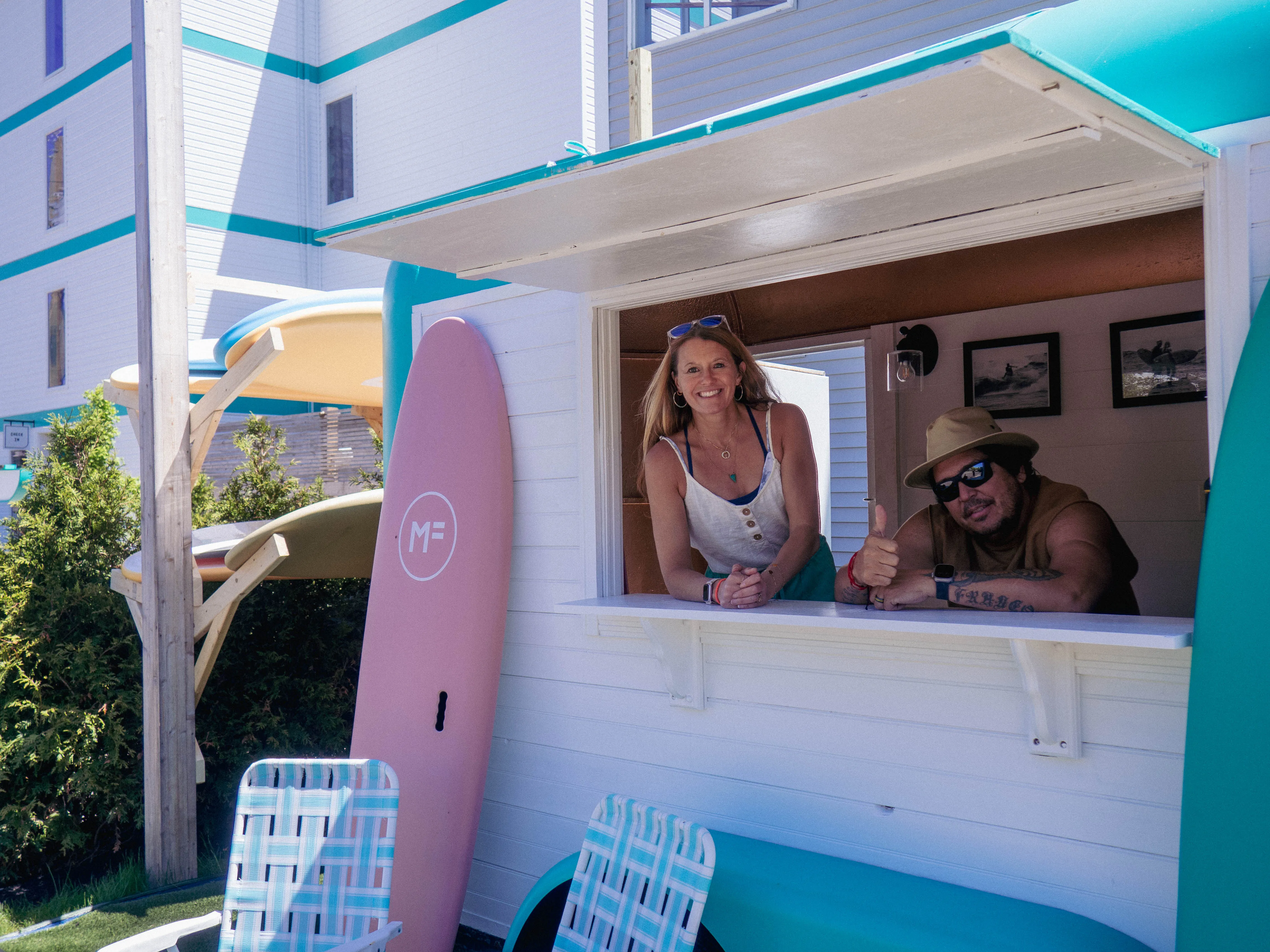A smiling woman and man leaning out of a white kiosk window with surfboards and lawn chairs nearby.