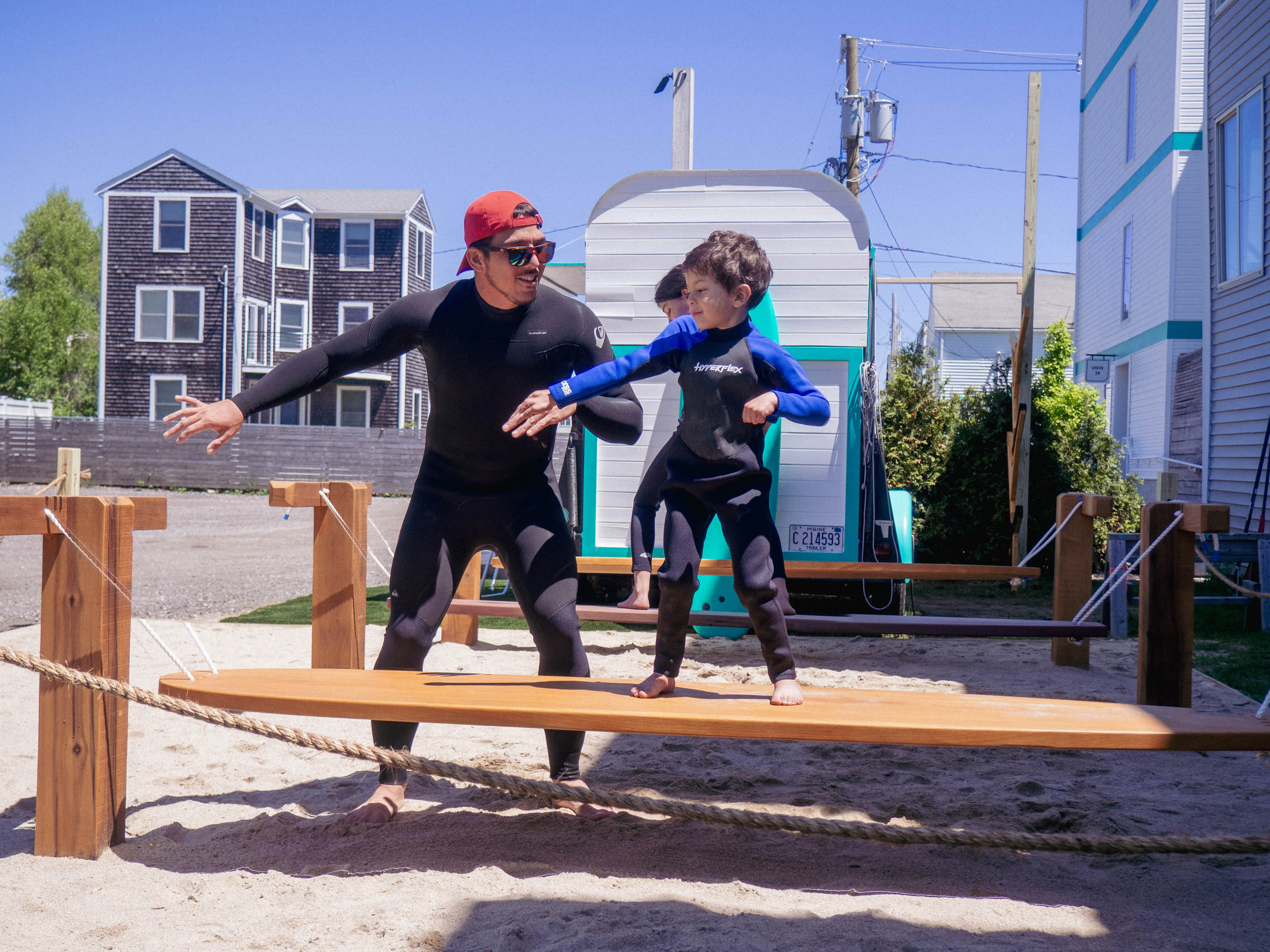 Man wearing wetsuit and red cap helping child balance on wooden plank in sandy outdoor playground.