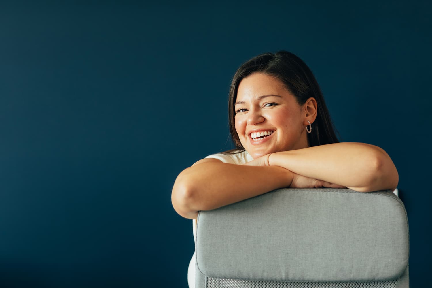 Femme souriante appuyée sur le dossier d'une chaise grise devant un mur bleu sombre.