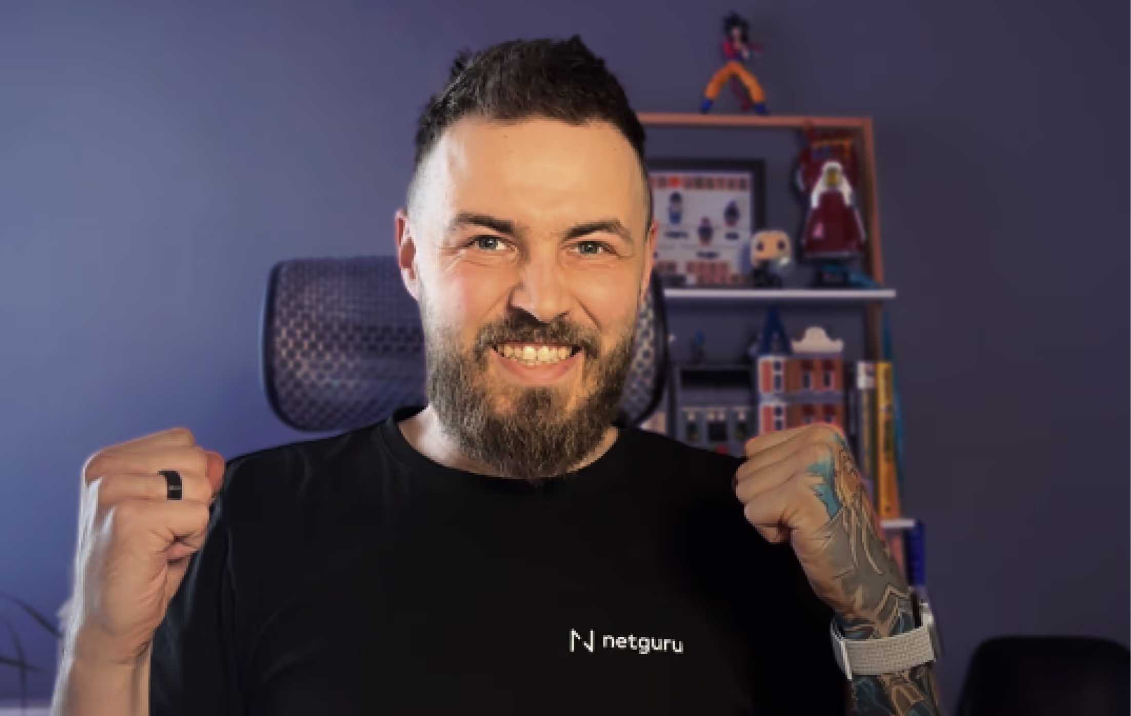 Smiling bearded man wearing a black netguru t-shirt raising both fists in excitement, with a dark purple background and shelves holding figures and books behind him.