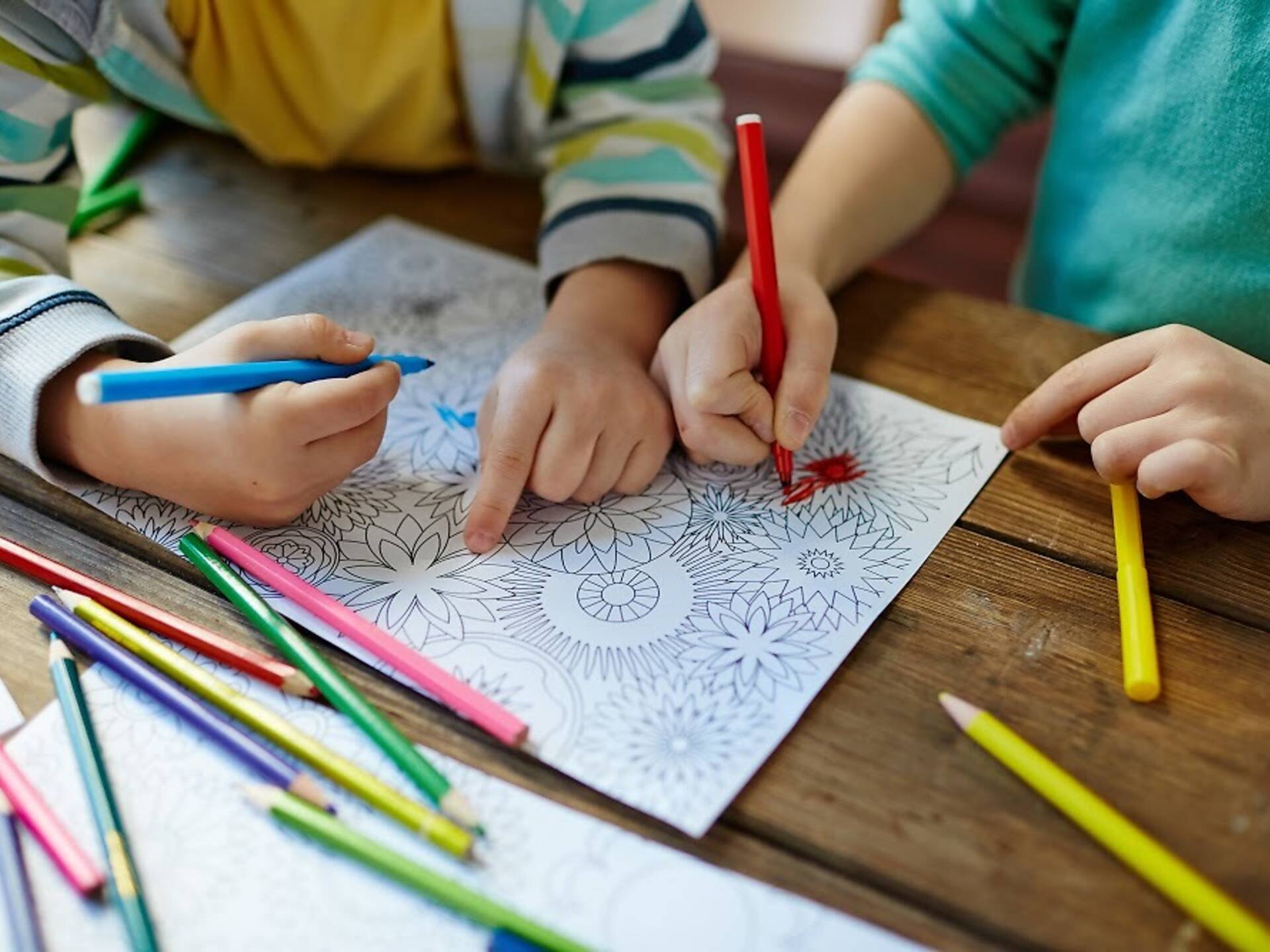 Two children coloring a floral-themed coloring page on a wooden table using colored pencils and markers, with one child coloring with a red marker and the other holding a blue marker.