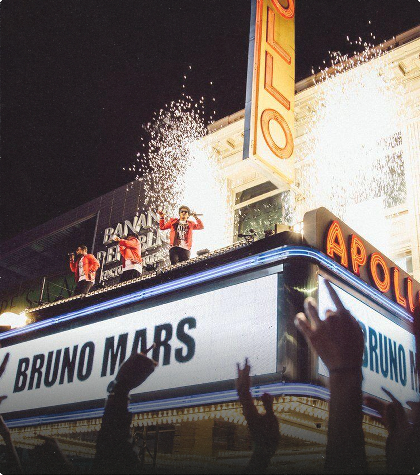Bruno Mars performing on top of the Apollo Theater marquee with fireworks and audience hands raised at night.