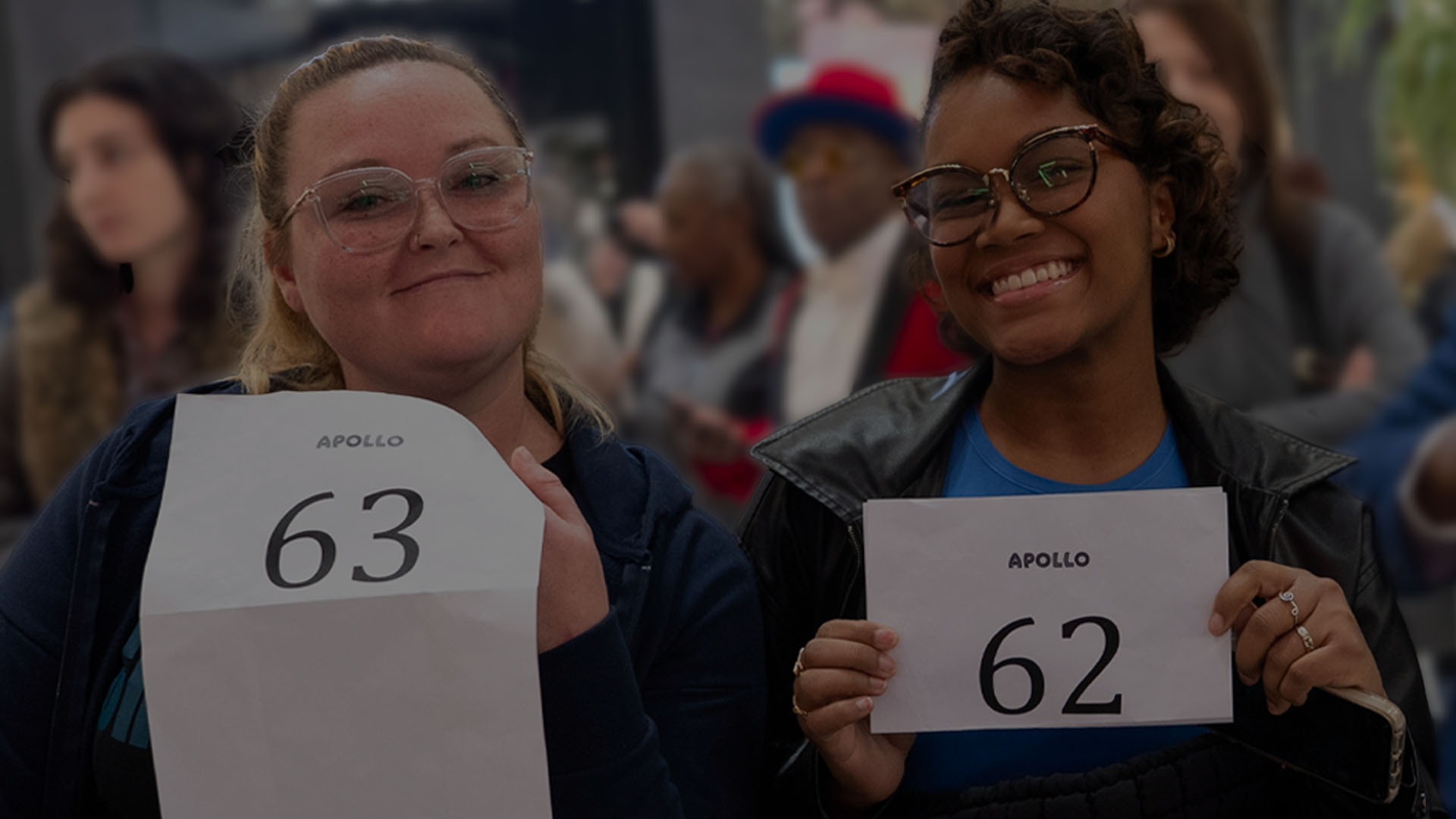 Two women holding Apollo audition numbers and smiling inside a crowded venue.