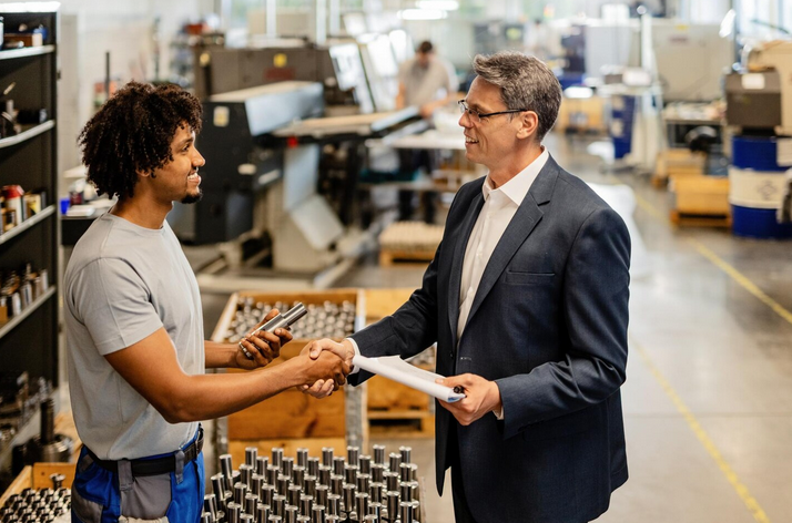 Smiling businessman in a suit shaking hands and greeting a steel worker wearing safety equipment in an industrial manufacturing facility, illustrating successful business relationships, sales partnerships, and client engagement in the manufacturing industry.
