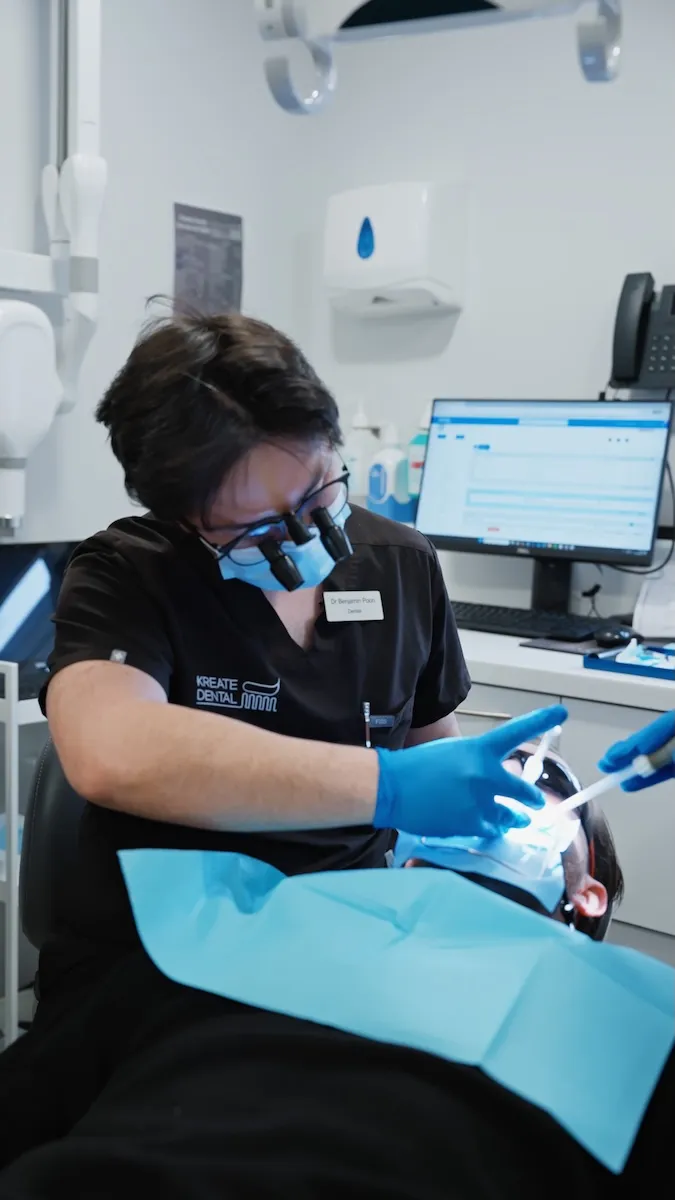 Dentist wearing magnifying glasses and mask treating patient in a dental clinic with tools and computer screen.