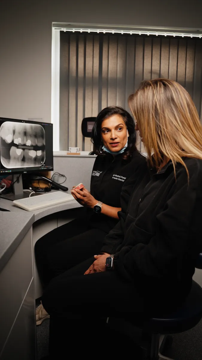 Dentist explains dental X-ray on computer to patient in an office with vertical blinds.