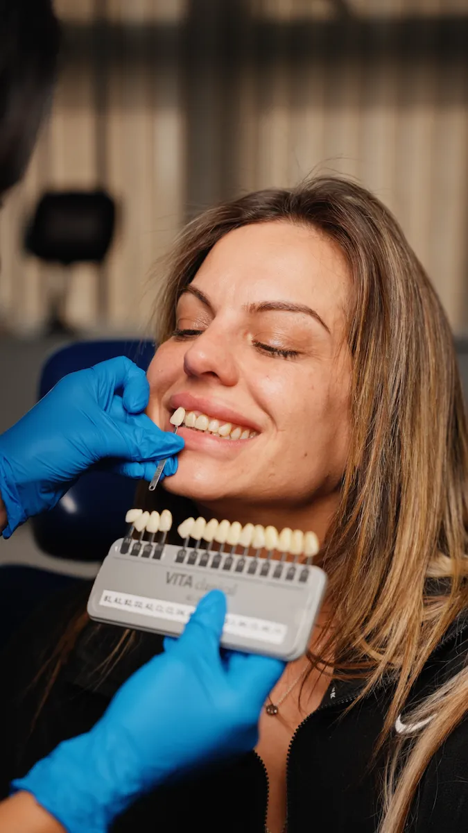 Dentist wearing blue gloves matching shade of dental crown to smiling woman's teeth.