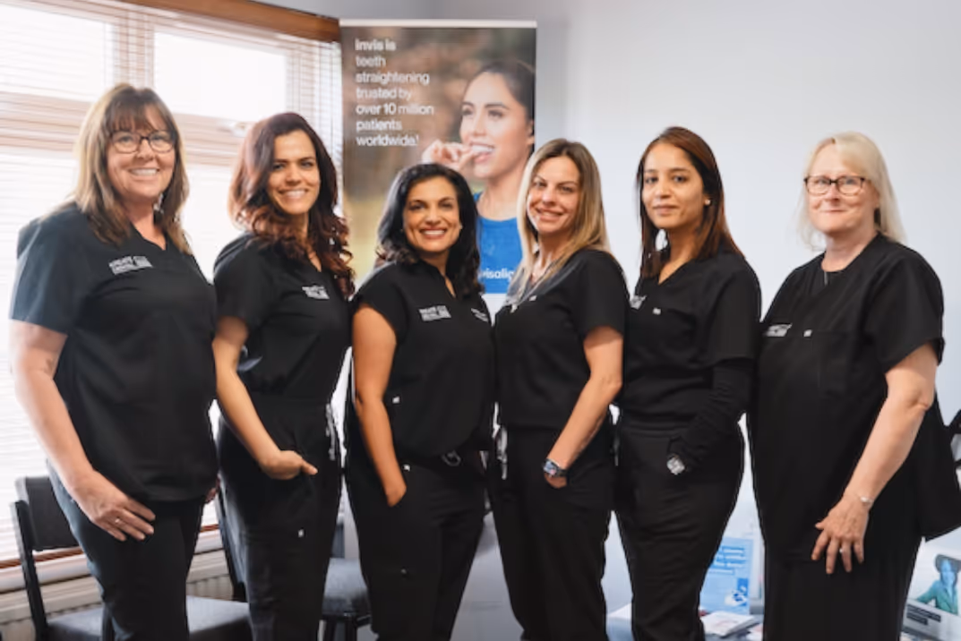 Six women in black uniforms standing indoors, smiling, with a poster about Invisalign behind them.