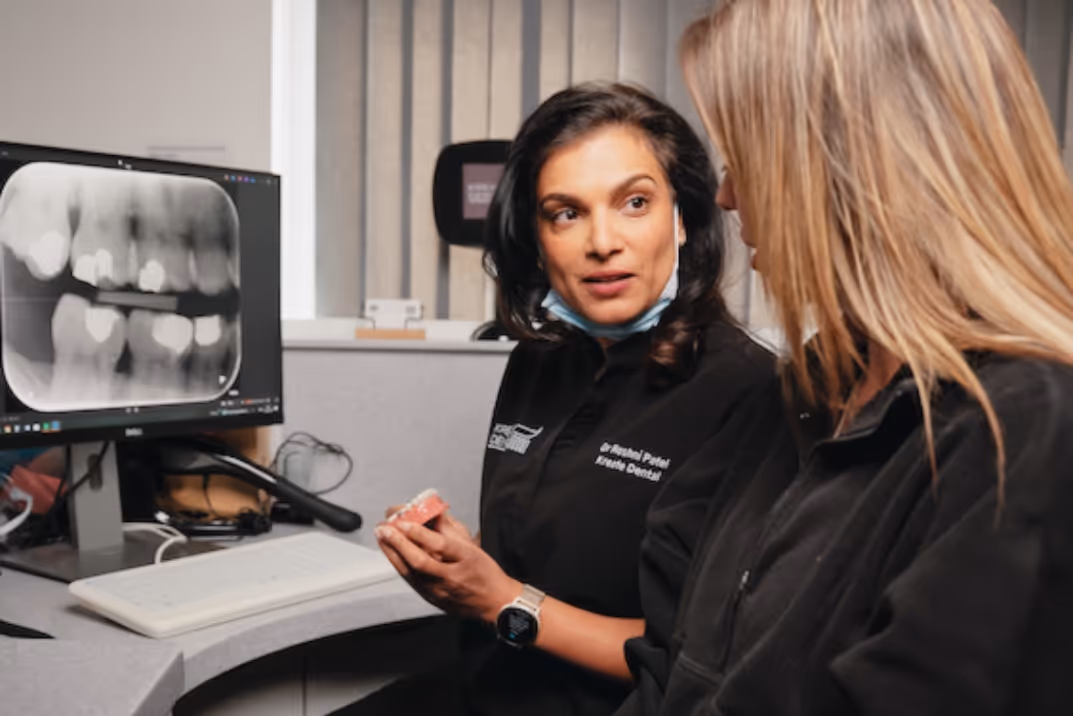 Female dentist explains dental model to patient with tooth X-ray visible on computer screen.